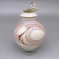 Polychrome lidded jar with a fine line and geometric design and a lid with an applique detail of a clay person and organic form sitting on a piece of wood
 by Carla Martinez of Mata Ortiz and Casas Grandes