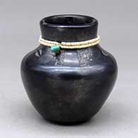 A black jar with a band of heishi beads around the neck with a piece of turquoise, above a band of sgraffito deer tracks and geometric design