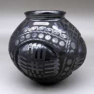 A glossy black jar with a flared rim, four repousse faces and decorated all over with a matte black geometric design
 by Eduardo Ortiz aka Chevo of Mata Ortiz and Casas Grandes