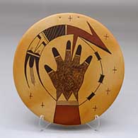 A round tile decorated with a hand, bird element and geometric design, plus holes for hanging
 by Garrett Maho of Hopi