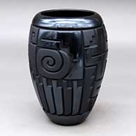 A matte and polished black cylinder decorated with a four-panel carved spiral, yei, feather and geometric design
 by Harrison Begay Jr of Santa Clara